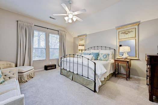 Bright and inviting primary bedroom featuring  plush carpeting and a ceiling fan. The room is accented with soft lighting, and ample natural light from two sizable windows. Cozy seating adds a touch of comfort.