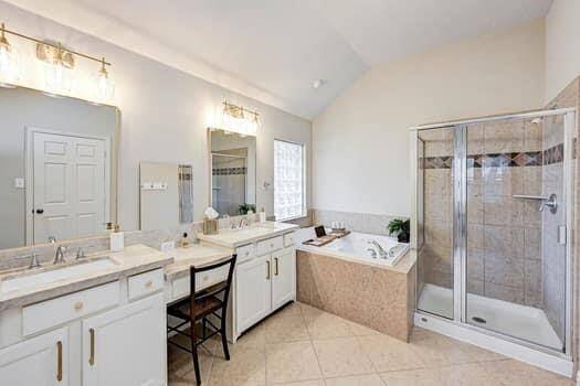 The primary bathroom features a spacious layout with dual sinks, a large vanity, and ample lighting. It includes a separate glass-enclosed shower and a luxurious corner bathtub with elegant tile accents. The neutral color palette and high ceiling add to the open, airy feel.