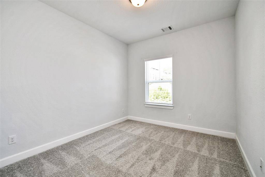 This is a bright, empty room with light gray walls and carpet. There's a single window letting in natural light, and the room has a ceiling light fixture, making it ideal for a small bedroom or office space.