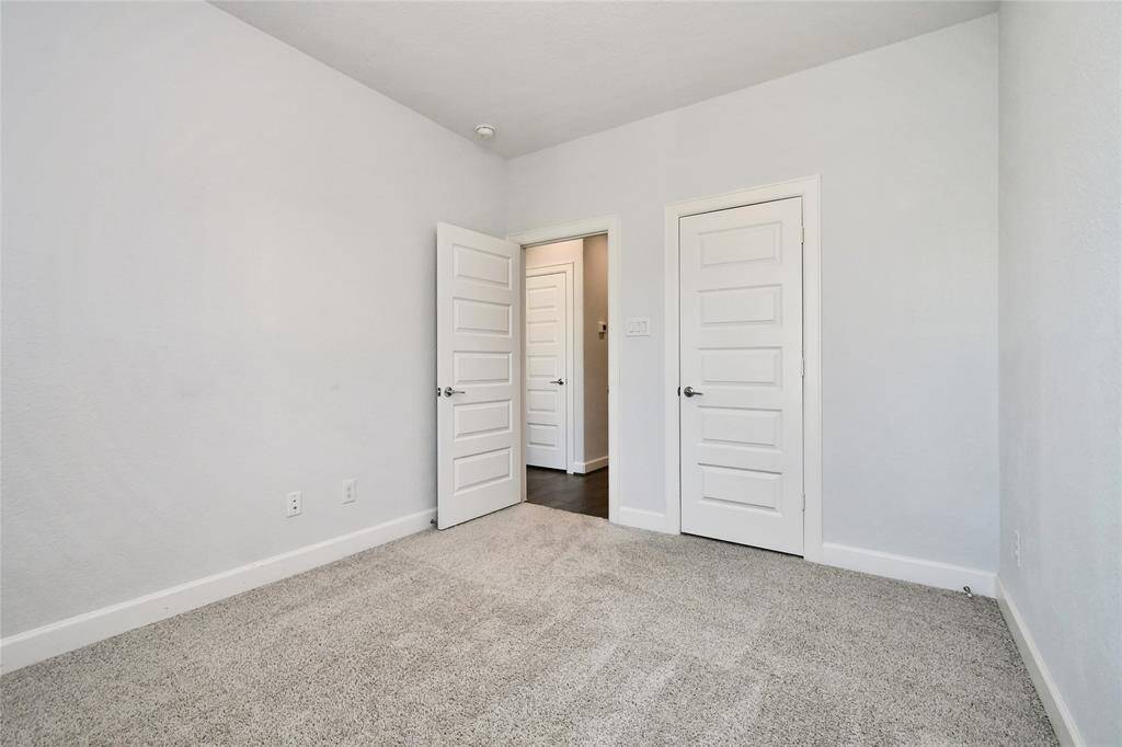 This is a bright, neutral-toned bedroom with carpeted flooring. It features two white paneled doors, one leading to a hallway. The space is simple and ready for personal touches.