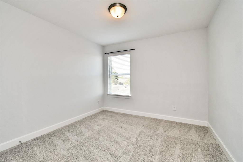 This photo shows a bright, empty room with light gray walls, plush carpet flooring, and a single window allowing natural light. The ceiling has a simple light fixture, creating a clean and neutral space ideal for customization.