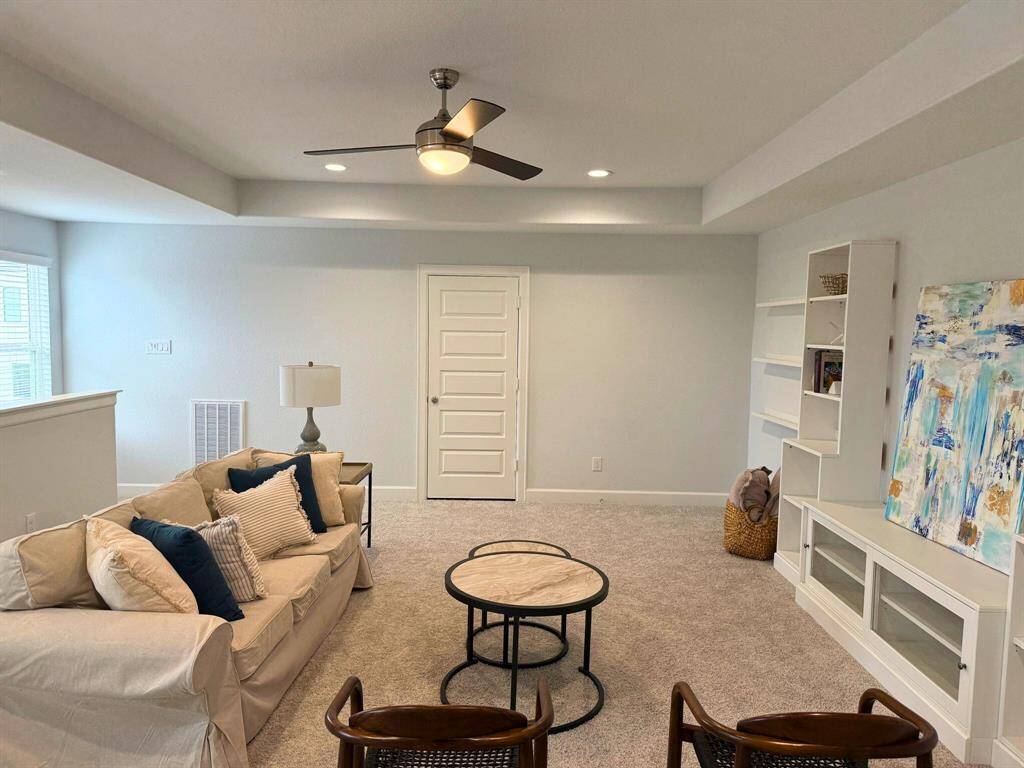 This cozy living area features a neutral-toned sofa with accent pillows, a modern ceiling fan, and a small round coffee table. Built-in shelves and a colorful abstract painting add character, while natural light streams in from the window. The space is carpeted, offering a warm and inviting feel.