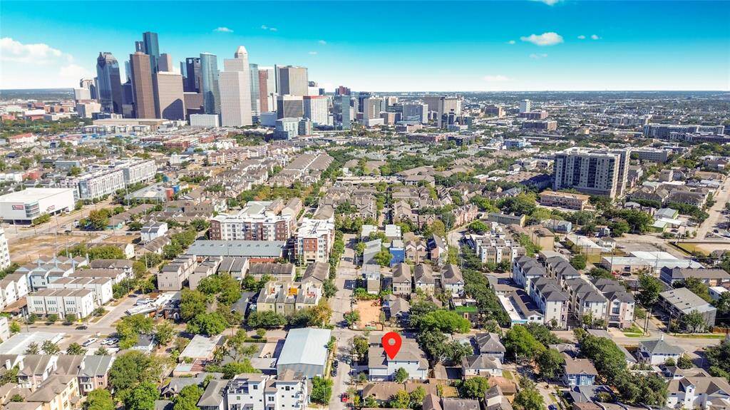 Aerial view of a residential neighborhood with a prominent red marker indicating a specific property. The area is surrounded by greenery and features a mix of homes and mid-rise buildings. In the background, a vibrant city skyline is visible, suggesting a convenient urban location.