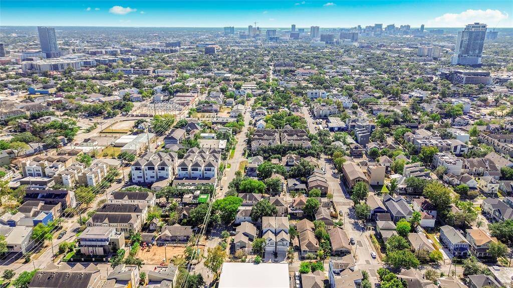 This aerial view showcases a vibrant urban neighborhood with a mix of residential homes and modern townhouses. The area is lush with greenery and offers a clear view of the city skyline in the distance, suggesting proximity to urban amenities and a lively community atmosphere.