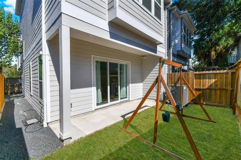 This photo showcases the backyard of a modern three-story home with gray siding. It features a small grassy area with a concrete patio adjacent to a sliding glass door. The space is enclosed by a wooden fence, offering privacy.