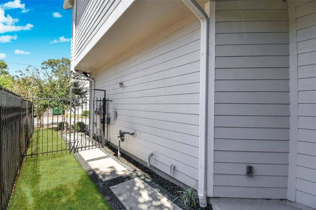 This image shows a narrow side yard with a walkway made of stepping stones alongside a house with light gray siding. A black metal fence encloses the area, and there are small plants along the edge. The area is well-maintained with a grassy strip.