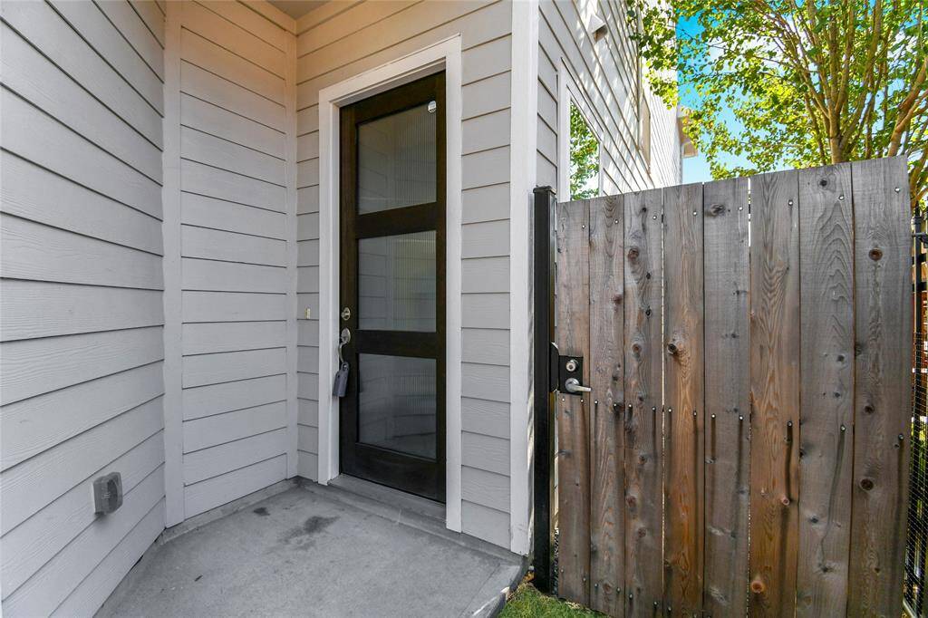 This photo shows a small, private entryway with a modern glass door and light gray siding. Adjacent is a wooden gate, providing privacy and a charming entrance to the home.