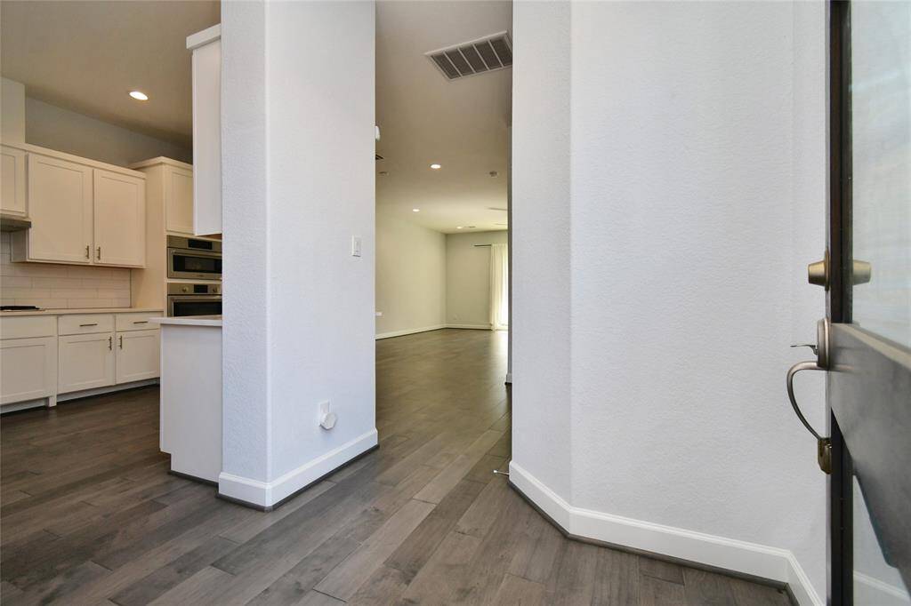 This photo shows a bright, modern entryway leading into an open-concept space with light-colored walls and dark wood flooring. The kitchen features white cabinetry and built-in stainless steel appliances, giving a sleek and contemporary feel.