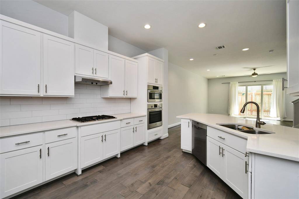 This photo showcases a modern kitchen with sleek white cabinetry, stainless steel appliances, and a large island with a sink. The space features recessed lighting, a gas cooktop, and ample natural light from nearby windows, offering a bright and open feel.