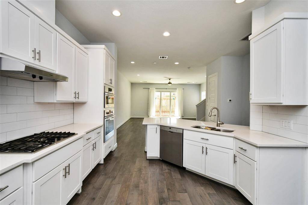 This modern, open-concept kitchen features sleek white cabinetry, stainless steel appliances, and a large island with a sink. It flows seamlessly into a bright living area with wood flooring, recessed lighting, and a view of the backyard through sliding glass doors.