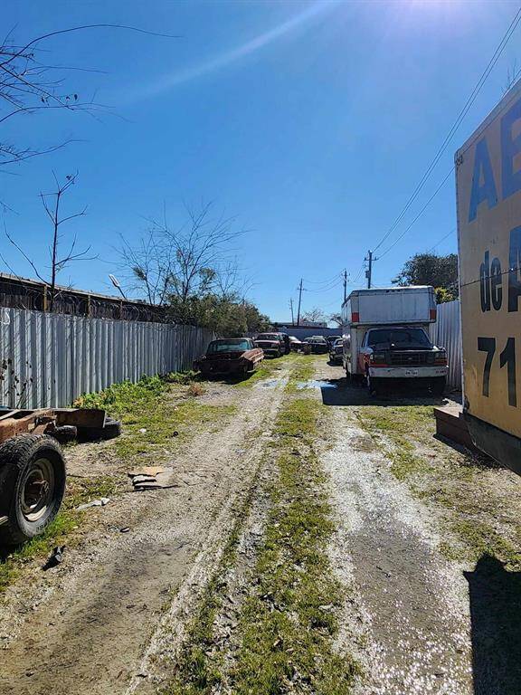 This photo shows a narrow, unpaved driveway lined with metal fencing and vehicles, suggesting a storage or industrial area. The ground is grassy with some muddy patches, and there are a few scattered pieces of debris.