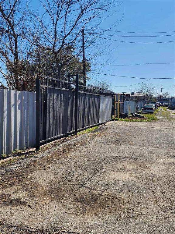 This photo shows a narrow alleyway with a paved surface and a metal fence on one side. The area appears to be a back access road for properties, with some parked vehicles visible in the distance. The surroundings suggest a quiet, urban environment.
