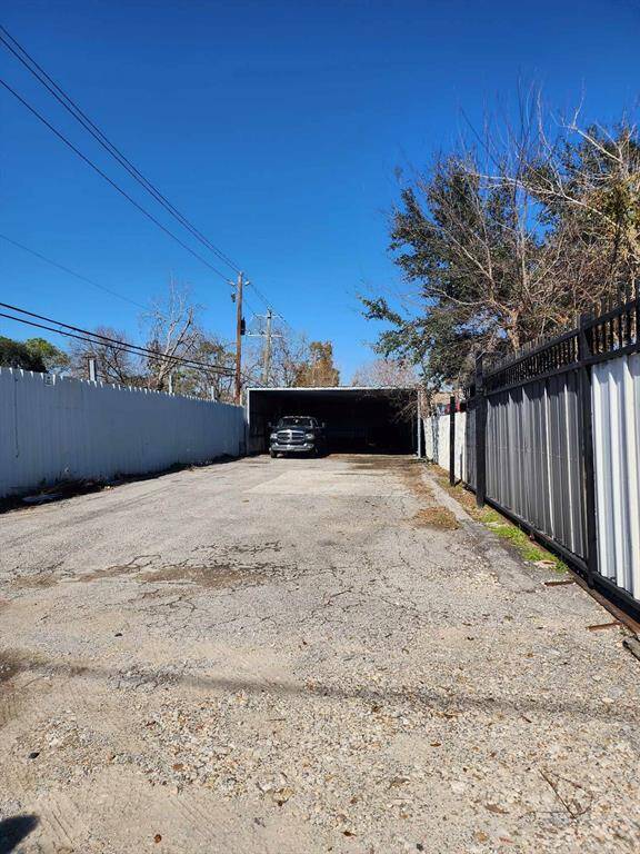 This image shows a long driveway with a carport at the end, providing covered parking for vehicles. The area is enclosed by fences on both sides, featuring a mix of paved and gravel surfaces. The setting is open to the sky with some trees visible.