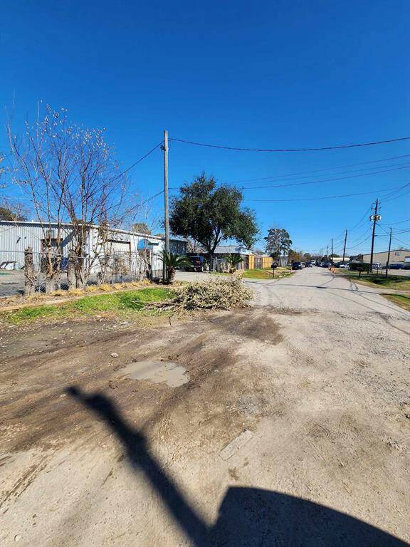 This photo shows a quiet residential street with a mix of paved and unpaved surfaces. There are utility poles and wires lining the street, alongside some trees and vegetation. The area appears to be calm and open, offering a rural or semi-industrial feel.