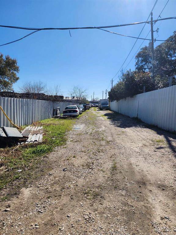 The photo shows a narrow, unpaved alley lined with cars and bordered by tall, corrugated metal fences. The area appears somewhat cluttered, with some patches of grass and debris along the sides. It’s a sunny day with clear skies.