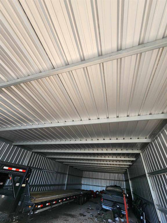 The photo shows a spacious metal storage unit or garage with a corrugated metal roof and walls. Inside, there is a large trailer and some equipment, indicating ample space for storage or vehicle parking.