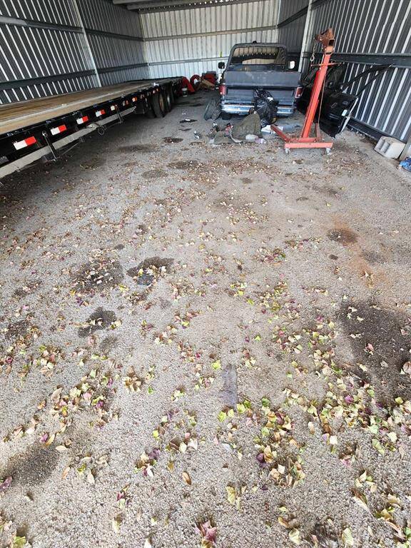 This photo shows a spacious metal storage unit or garage with gravel flooring. It's currently used to store a truck, trailer, and various equipment, with some scattered leaves and debris on the ground. Ideal for vehicle or equipment storage.