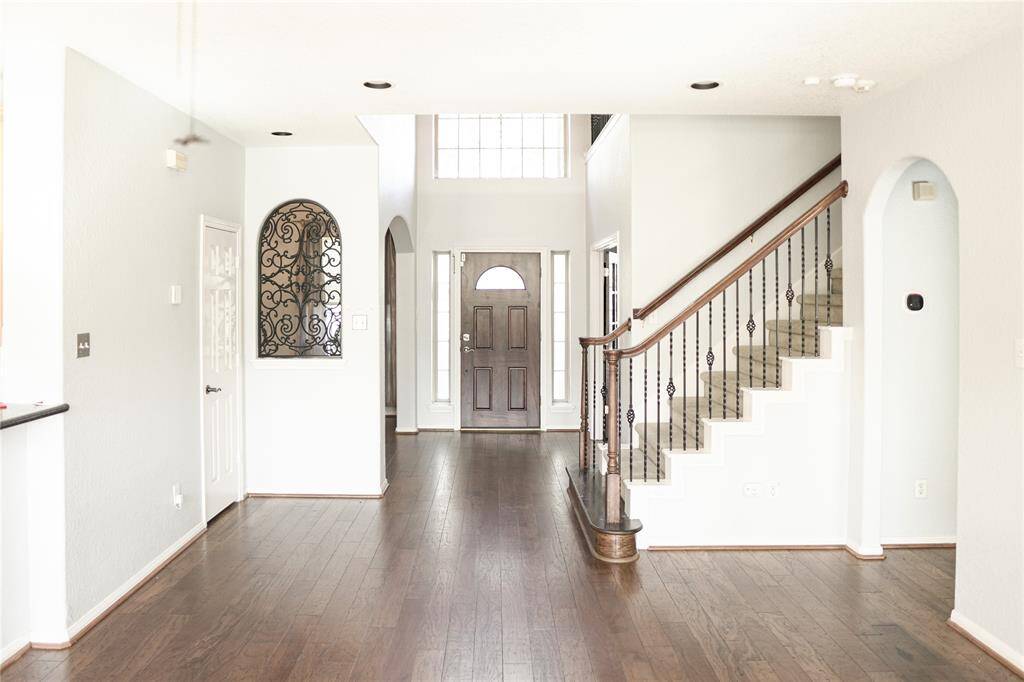 This welcoming entryway features hardwood floors, a stylish staircase with wrought iron balusters, and ample natural light from large windows. The space includes a decorative wall niche and a dark wood front door, offering a blend of elegance and functionality.