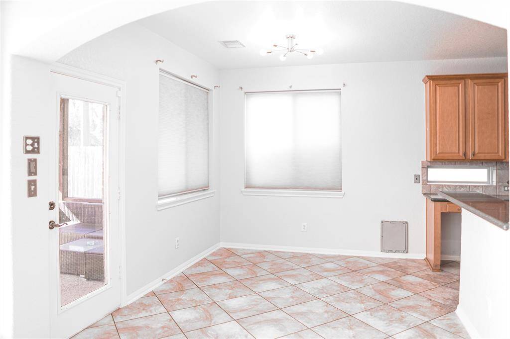 This bright room features tiled flooring and neutral walls, with ample natural light from two windows and a glass door leading outside. A modern light fixture and wooden cabinets provide a touch of style, while the open layout offers versatility.