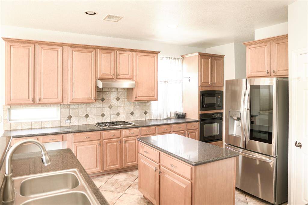 This kitchen features warm wood cabinetry, granite countertops, and stainless steel appliances, including a double oven and French-door refrigerator. A central island and dual sinks provide ample workspace, complemented by a stylish tile backsplash.