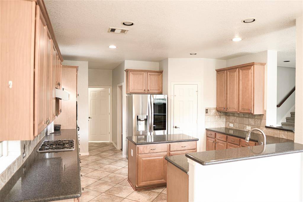 Spacious kitchen with light wood cabinetry, granite countertops, and stainless steel appliances. Features a central island, ample storage, and tiled flooring. Bright and open layout with access to adjacent areas.