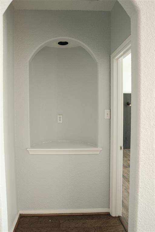This photo shows a small alcove with an arched top, painted in a light gray color. The space features a built-in shelf and an electrical outlet, ideal for a decorative display or small appliance. Adjacent is a doorway leading to a room with wood flooring.