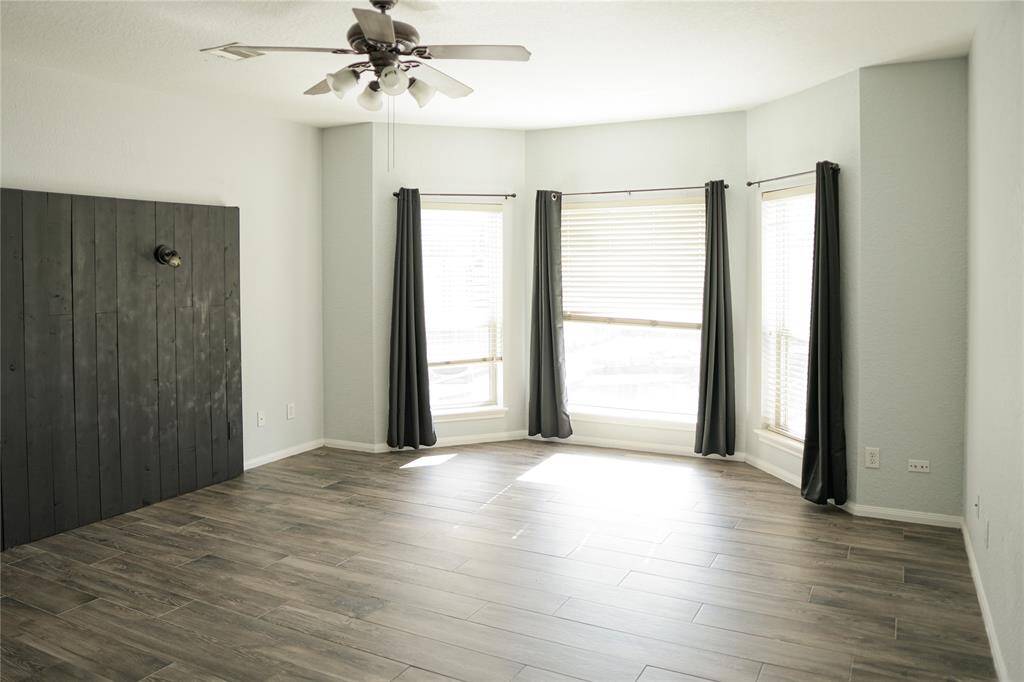 This room features a spacious area with light gray walls and wood-look tile flooring. It has a ceiling fan, a large bay window with blinds and dark curtains, providing ample natural light, and a unique dark wood accent wall.