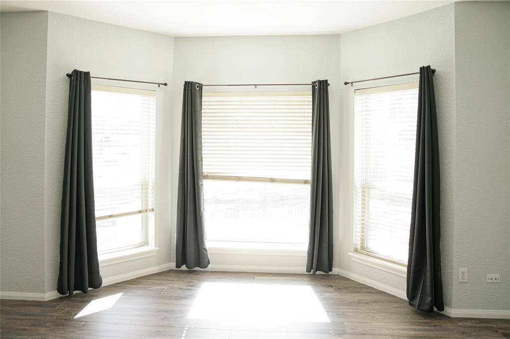 Bright room featuring large bay windows with blinds and dark curtains, complemented by light-colored walls and wood-look flooring. Ideal for natural lighting.