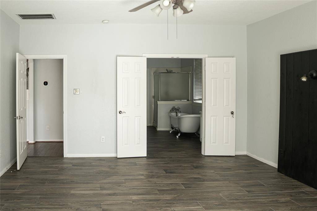 This photo showcases a spacious room with wood-look tile flooring and light gray walls. It features a ceiling fan and double doors leading to a bathroom with a standalone tub. The room has a modern, minimalistic design with ample natural light.