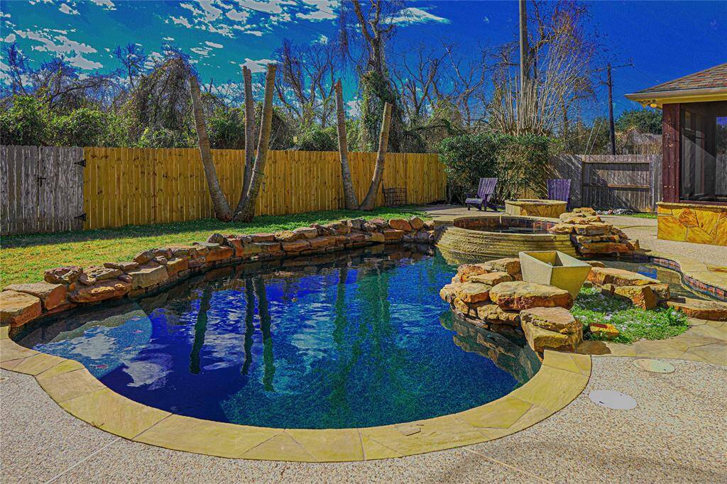 The photo shows a backyard with a stylish in-ground pool surrounded by natural stone edging. There's a cozy seating area and a hot tub, offering a serene, private escape with tall fencing and lush greenery. Perfect for relaxation and entertaining.