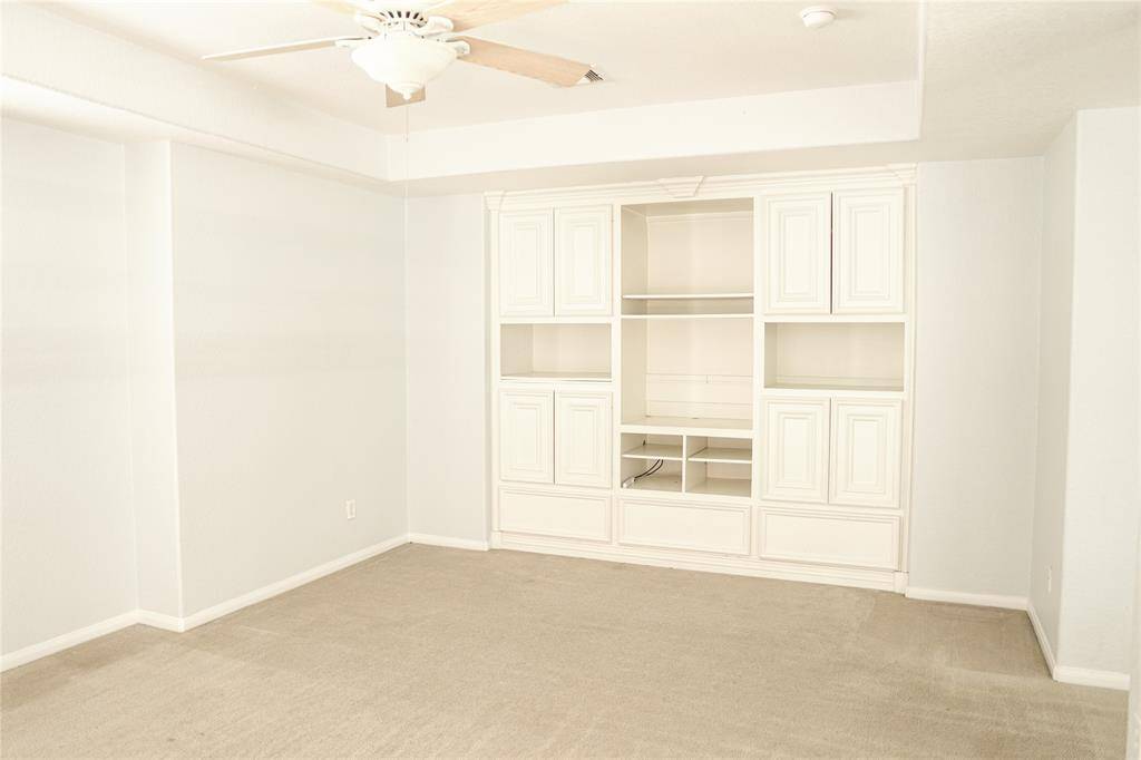 This room features a built-in white entertainment center with ample shelving and cabinets, neutral carpeting, and a ceiling fan. The light-colored walls create a bright and inviting space.
