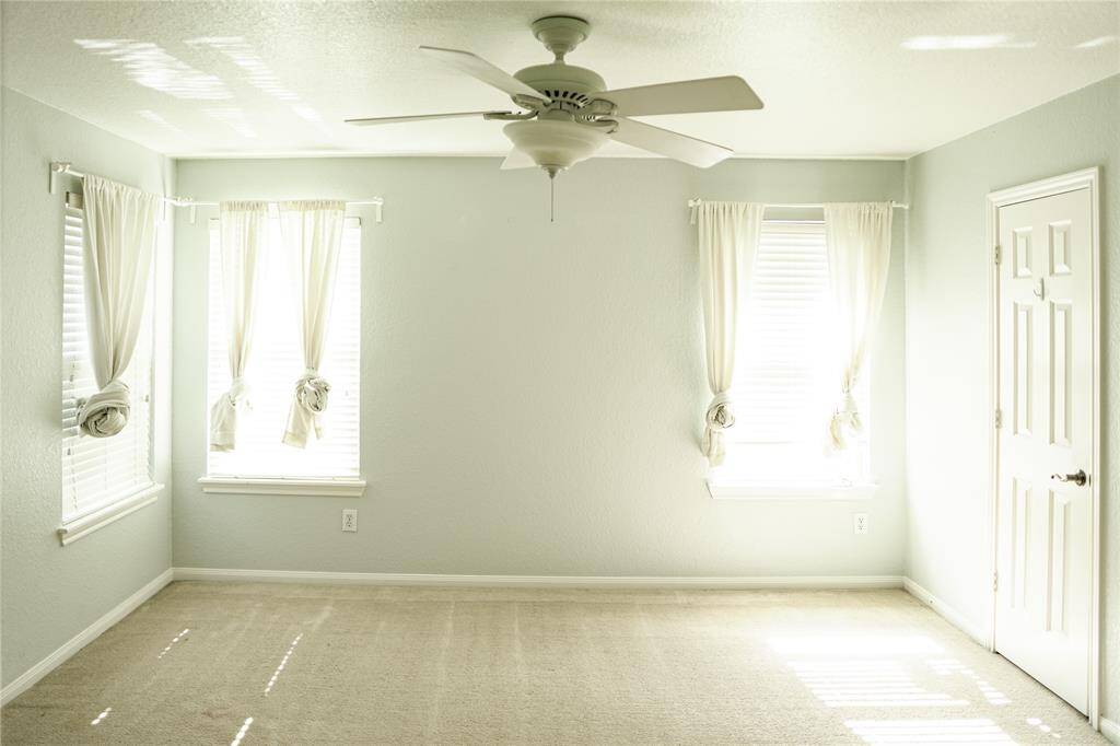 Bright, spacious room with neutral walls and carpet. Features two large windows with sheer curtains, letting in plenty of natural light. A ceiling fan is centrally located, and there's a white door on the right.