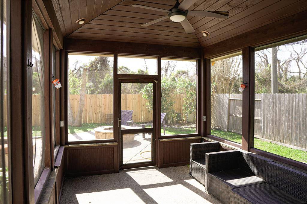 This photo shows a cozy screened-in porch with wooden paneling and a ceiling fan, offering a view of a fenced backyard with a lawn and a fire pit area. It's a perfect spot for relaxation and outdoor enjoyment.