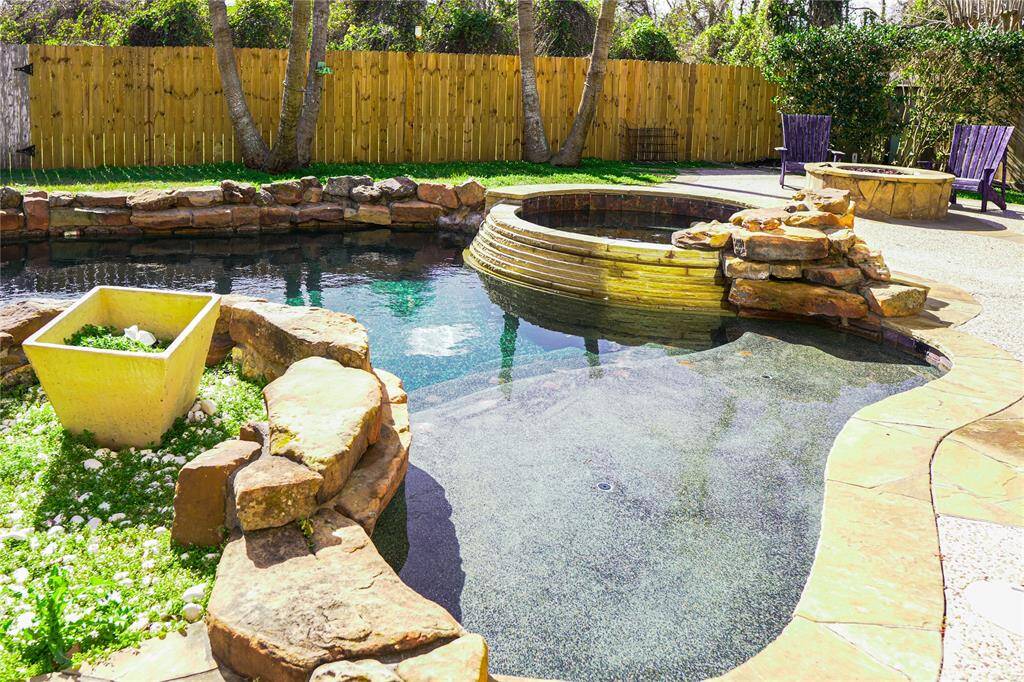 This backyard features a beautifully landscaped pool and spa, surrounded by natural stone accents. There is a wooden fence for privacy, a manicured lawn, and a cozy seating area with a fire pit, perfect for outdoor relaxation and entertaining.