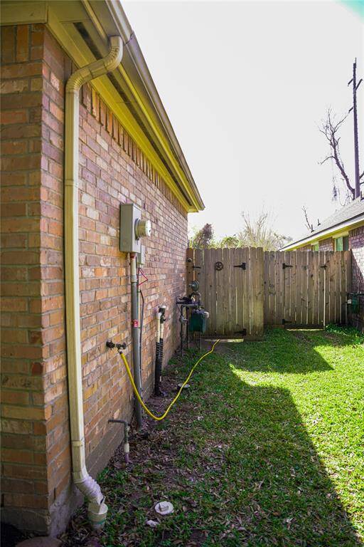 This photo shows a narrow side yard with a brick exterior wall, utility connections, and a wooden privacy fence. The area has a grassy lawn, suitable for light outdoor activities or gardening.