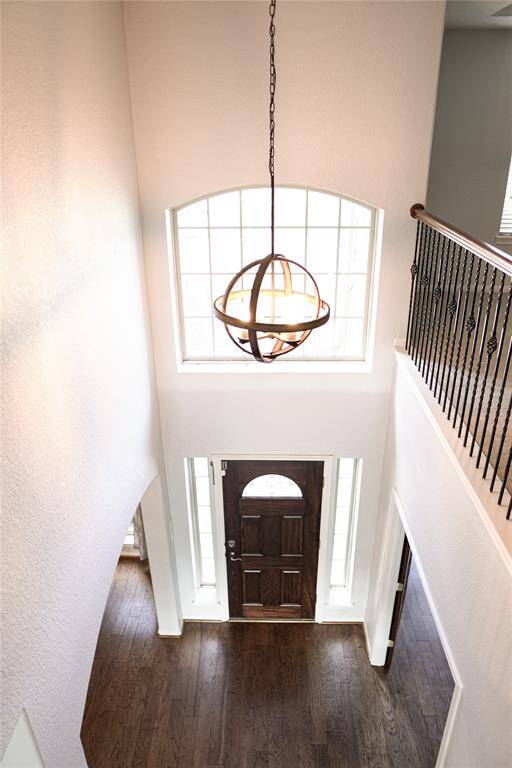 This photo showcases a bright, spacious foyer with a high ceiling and large window, allowing plenty of natural light. A stylish pendant light hangs overhead, and there's a wooden front door with sidelights, complemented by dark hardwood flooring and elegant wrought iron railing.