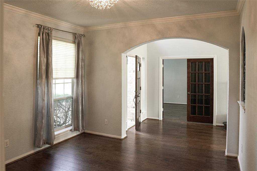 This photo showcases a bright entryway with hardwood floors, a large window with curtains, and an elegant archway leading to a room with French doors. The space is enhanced by crown molding and a modern light fixture, offering a welcoming ambiance.