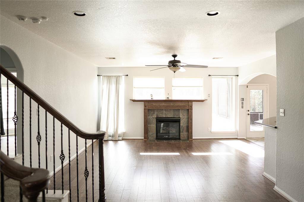 This photo showcases a bright living room featuring a ceiling fan, a tiled fireplace, and large windows that let in ample natural light. The space has hardwood floors and opens to an adjacent area with a door leading outside. A staircase with wrought iron railings adds elegance to the room.