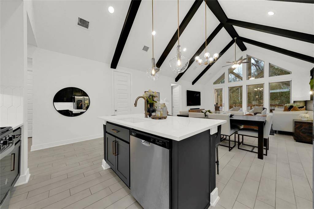 This open-concept kitchen and living area features vaulted ceilings with exposed beams, modern pendant lighting, and large windows for natural light. The kitchen island space flows seamlessly into the dining and comfortable area, perfect for entertaining.