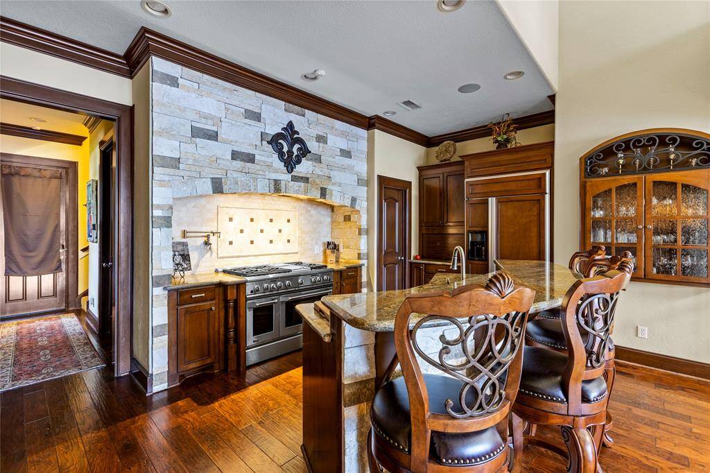 This is a spacious kitchen featuring rich wooden cabinetry, a stone backsplash, granite countertops, and a professional-grade GE Monogram over and range. A central island with bar seating and elegant ironwork detailing adds to the charm, while the hardwood floors give it a warm, inviting feel.