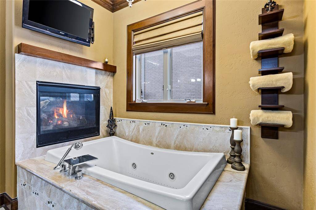 This is a luxurious bathroom featuring a large jetted tub with a marble surround, a cozy, double-sided fireplace, and a mounted flat-screen TV. It has a large window providing natural light and a stylish towel rack on the wall.