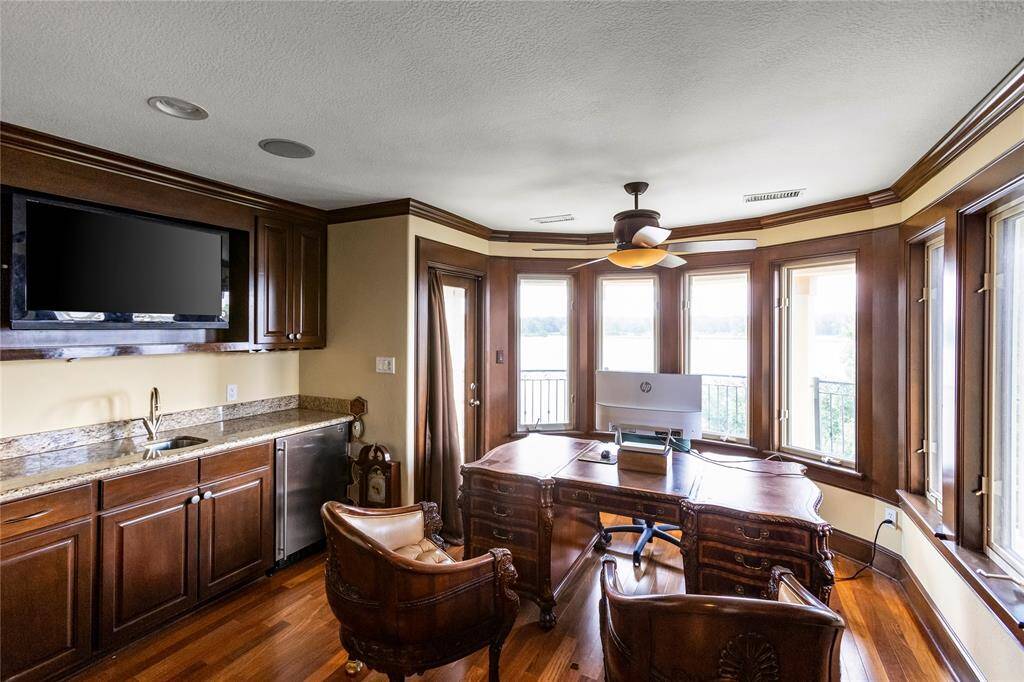 The photo showcases a well-appointed home office with rich wooden cabinetry, a built-in desk area, and ample natural light from large windows offering an aerial view of Lake Conroe. The room is equipped with modern amenities such as a flat-screen TV  and wet bar!