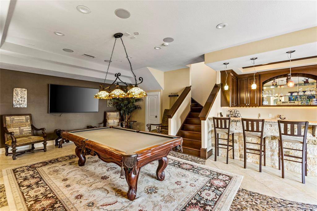This is a spacious, multi-purpose room featuring a pool table, a comfortable seating area, and an elegant bar with seating. The room is well-lit with a combination of pendant lights and a chandelier, and there's a large flat-screen TV mounted on the wall. A beautiful staircase leads to the upper level, showcasing the home's open and inviting layout.