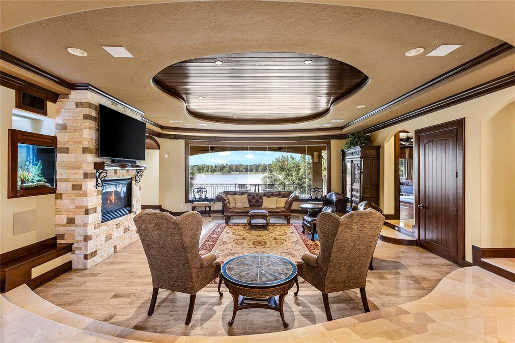This is an elegant living room with a stone fireplace, mounted flat-screen TV, and a coffered ceiling. It features marble floors and space for comfortable seating, with large windows and doors leading to a balcony with Lake Conroe views.