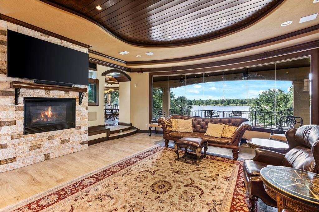 This is a luxurious living space featuring a cozy fireplace with stone surrounded, dual fish tanks and elegant wood-paneled ceilings. Expansive glass windows offer a stunning view of Lake Conroe. Traditional furniture complements the room's warm and inviting ambiance.
