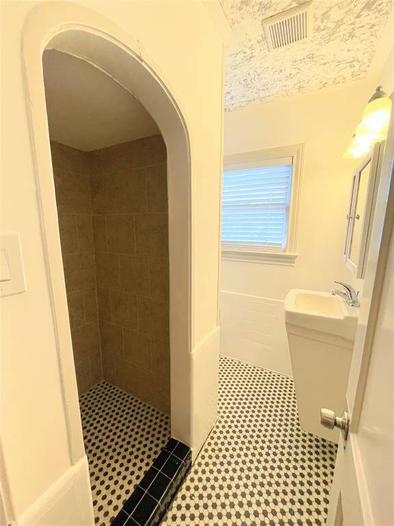 This en-suite features a tiled walk-in shower with an arched entrance, a compact rectangular sink, and a window for natural light. The floor has a classic black and white patterned tile.