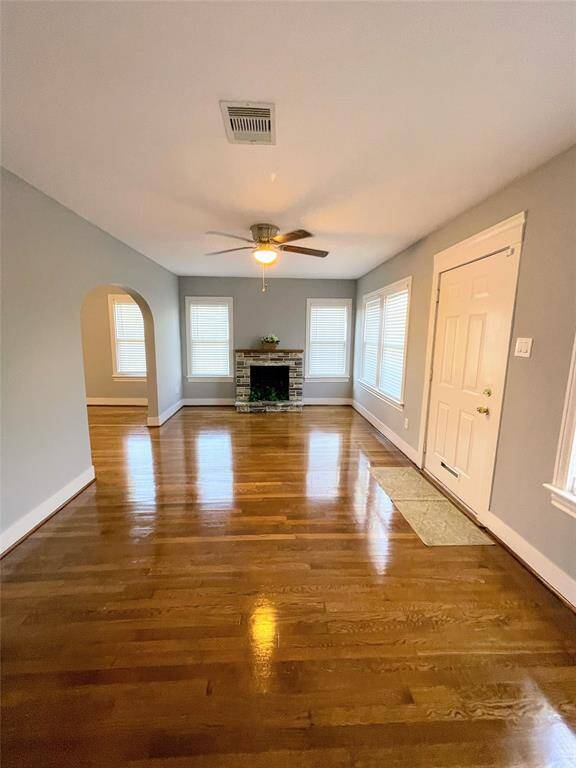 This photo showcases a bright living room with polished hardwood floors, a ceiling fan, and a central fireplace. The space has an arched entryway and multiple windows for natural light.