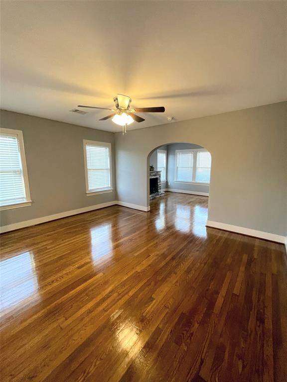 Spacious family room with polished hardwood floors, neutral walls, and a ceiling fan. It features ample natural light from the windows and an archway leading to a formal living room.