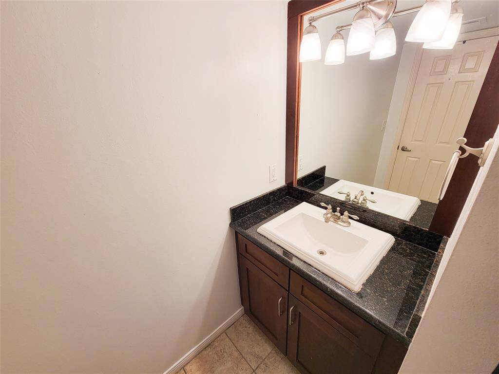 Dark wood vanity features a black granite countertop with decorative overmount sink. Above the vanity is a large mirror with an updated lighting fixture. The room has neutral wall colors and tile flooring.