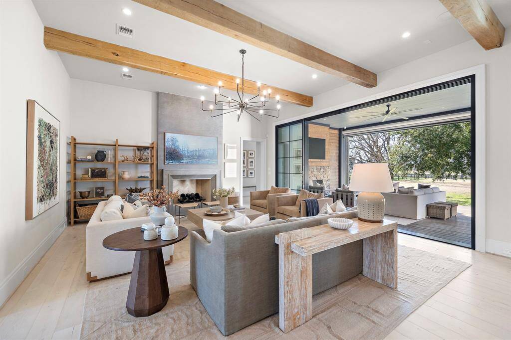 This spacious living room features exposed wooden beams, a modern chandelier, and a cozy fireplace. It seamlessly connects to an outdoor patio through large sliding glass doors, offering a picturesque view.